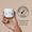 En hand håller en vit burk med Organic Babies Lugnande Salva för Bebis bredvid en Dermatologically Tested-logotyp på en beige bakgrund.